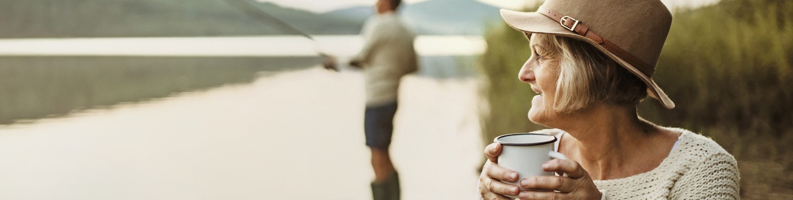 A woman with a tea mug and a man fishing by a lake